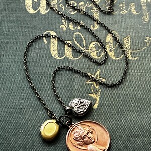 May include: A necklace with a black chain and three charms: a gold oval locket, a silver heart, and a copper medallion with a woman's face.