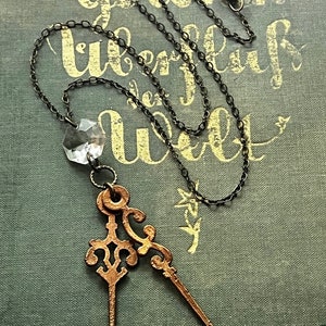 Puede incluir: Un collar delicado con un colgante de cristal y dos agujas de reloj de bronce de estilo antiguo. El collar está sobre un libro verde oscuro con letras doradas que dicen "vom gold'nen uberfluß der welt".