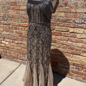 May include: A floor-length, embellished gown in shades of brown and silver. The dress features a square neckline, a fitted bodice with vertical and geometric patterns, and a flowing skirt with a layered, flared hem. The dress is displayed on a mannequin.