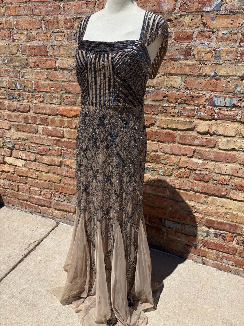 May include: A floor-length, embellished gown in shades of brown and silver. The dress features a square neckline, a fitted bodice with vertical and geometric patterns, and a flowing skirt with a layered, flared hem. The dress is displayed on a mannequin.