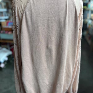Puede incluir: Un su&eacute;ter de color melocot&oacute;n claro con mangas largas. Las mangas tienen un panel transparente en el hombro y est&aacute;n adornadas con peque&ntilde;os tachuelas plateadas dispersas. El su&eacute;ter tiene un cuello y pu&ntilde;os acanalados.