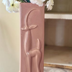 May include: A tall, rectangular, dusty rose-colored vase with a raised relief of a deer. The vase is filled with pink and white flowers and green leaves. The vase is sitting on a wooden surface.
