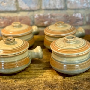 May include: Four ceramic soup bowls with lids. The bowls are tan with orange and blue stripes and a handle on the side. The lids have a small knob on top. The bowls are on a wooden surface.