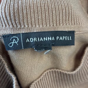 Puede incluir: Primer plano de una prenda de punto de color beige. La etiqueta dice "ADRIANNA PAPELL" en texto blanco sobre un fondo negro. La prenda tiene una textura acanalada.