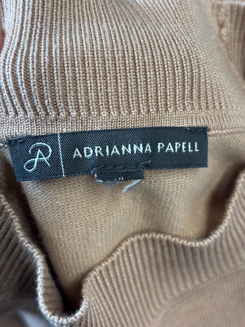 Puede incluir: Primer plano de una prenda de punto de color beige. La etiqueta dice "ADRIANNA PAPELL" en texto blanco sobre un fondo negro. La prenda tiene una textura acanalada.