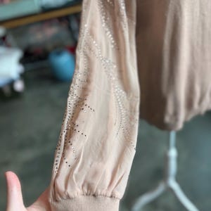 Puede incluir: Un su&eacute;ter de color melocot&oacute;n claro con mangas transparentes. Las mangas est&aacute;n adornadas con un patr&oacute;n de peque&ntilde;os adornos brillantes. El su&eacute;ter tiene un pu&ntilde;o acanalado y un cuello redondo.