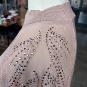 Puede incluir: Un su&eacute;ter rosa claro con cuello alto y paneles transparentes en los hombros. Los paneles transparentes est&aacute;n adornados con numerosas piedras de estr&aacute;s peque&ntilde;as y brillantes. El su&eacute;ter se exhibe en un maniqu&iacute;.
