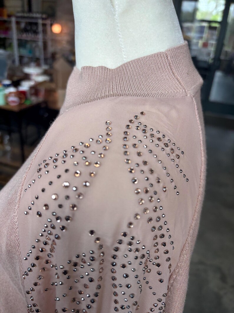 Puede incluir: Un su&eacute;ter rosa claro con cuello alto y paneles transparentes en los hombros. Los paneles transparentes est&aacute;n adornados con numerosas piedras de estr&aacute;s peque&ntilde;as y brillantes. El su&eacute;ter se exhibe en un maniqu&iacute;.