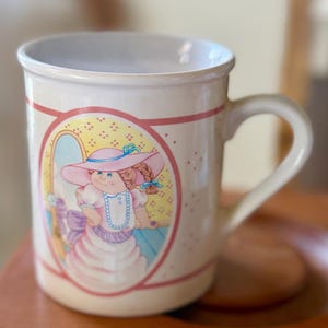 Puede incluir: Taza de cerámica color crema con una ilustración vintage de dos niñas con sombreros y vestidos. La taza tiene un borde rosa y amarillo alrededor de la ilustración y un asa curva. Un objeto de colección encantador.