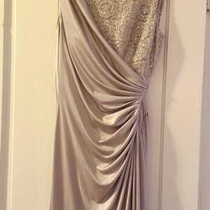 Puede incluir: Un vestido formal largo hasta el suelo en un suave color malva apagado. El vestido presenta un dise&ntilde;o asim&eacute;trico drapeado con un lado fruncido y un corpi&ntilde;o detallado y adornado. Cuelga de una percha de madera contra una puerta blanca.