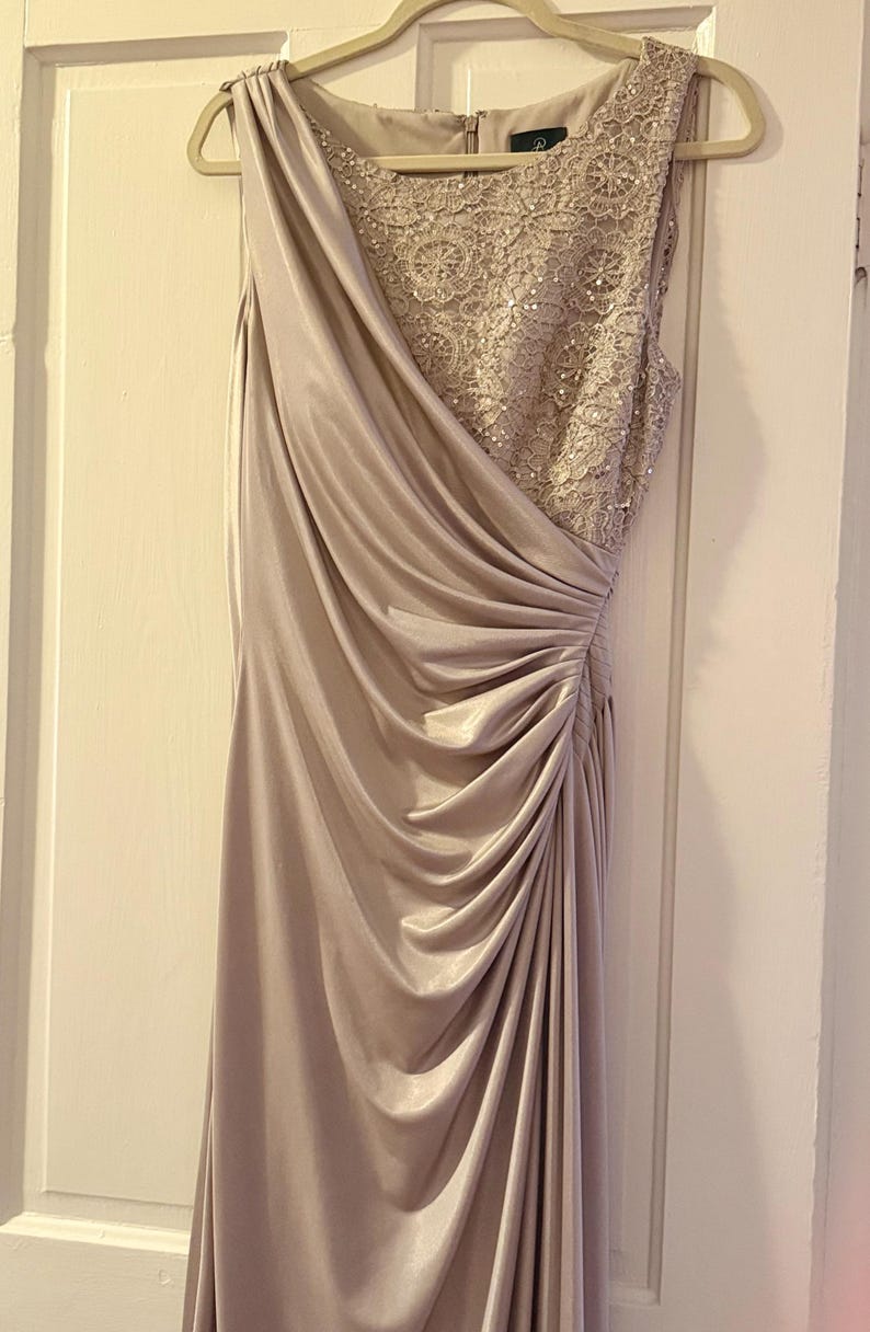 Puede incluir: Un vestido formal largo hasta el suelo en un suave color malva apagado. El vestido presenta un dise&ntilde;o asim&eacute;trico drapeado con un lado fruncido y un corpi&ntilde;o detallado y adornado. Cuelga de una percha de madera contra una puerta blanca.