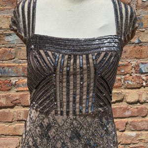 May include: A beige evening dress with intricate silver sequin and bead embellishments. The dress features a square neckline, cap sleeves, and a fitted bodice that flows into a detailed skirt. The dress is displayed on a mannequin against a brick wall.