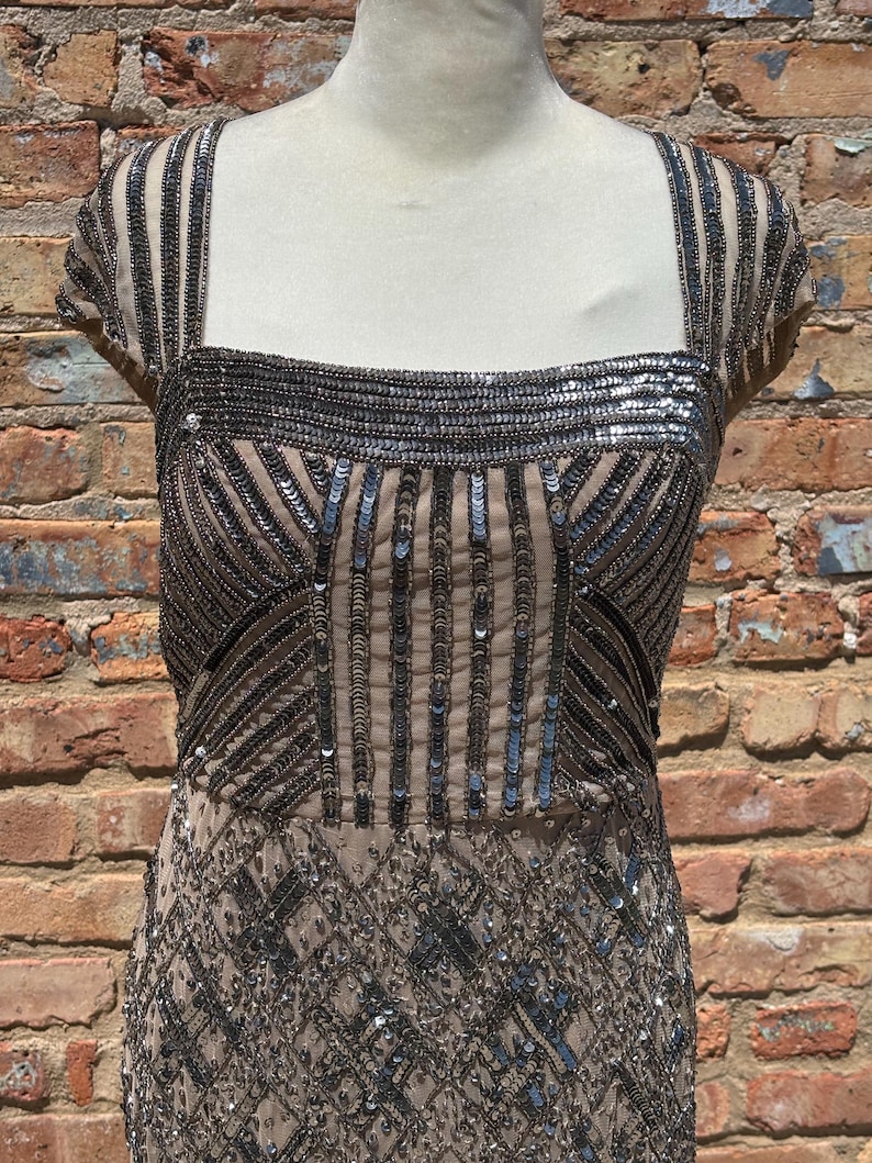 May include: A beige evening dress with intricate silver sequin and bead embellishments. The dress features a square neckline, cap sleeves, and a fitted bodice that flows into a detailed skirt. The dress is displayed on a mannequin against a brick wall.