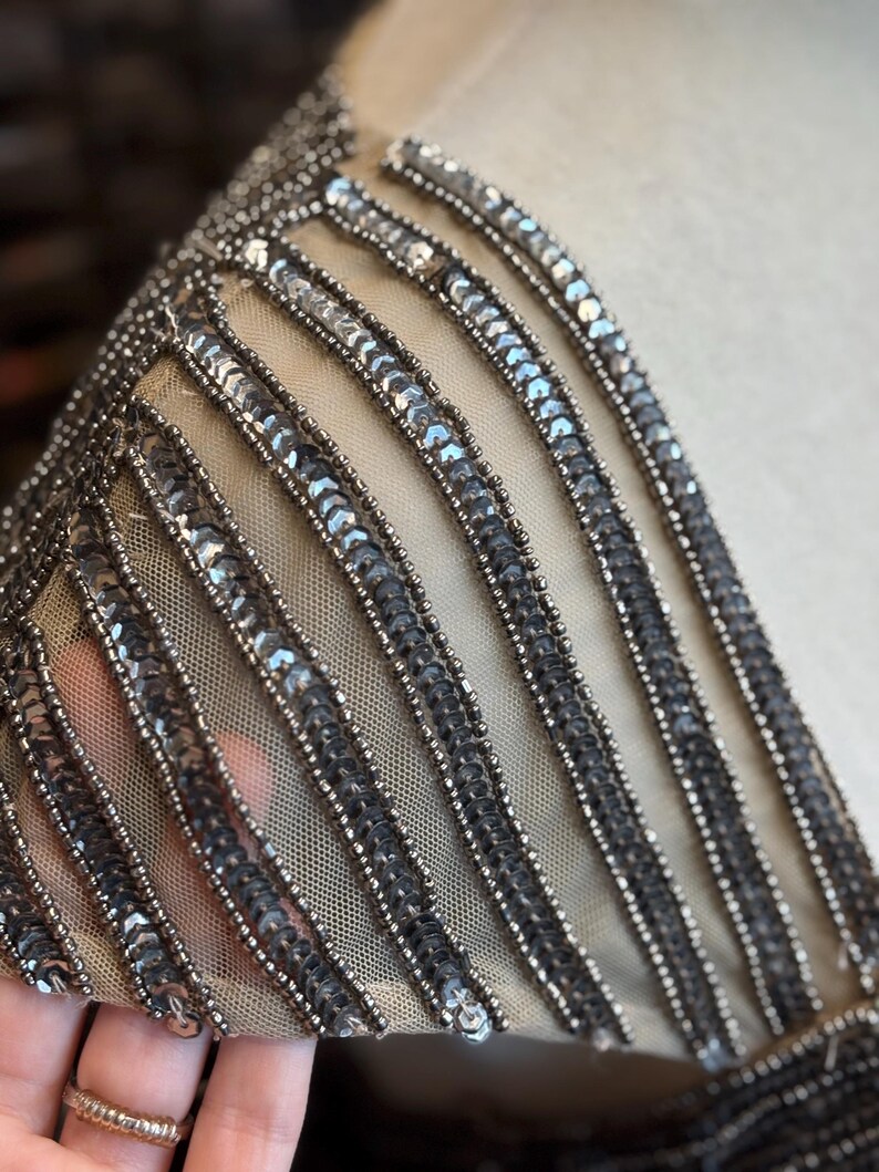 May include: Close-up of a section of a garment with intricate bead and sequin embellishments. The design features vertical rows of silver sequins and beads on a sheer, beige fabric, creating a shimmering effect. The details suggest formal wear.