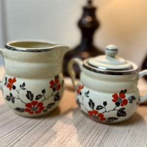 Puede incluir: Juego de azucarero y jarra de cerámica color crema. Ambas piezas presentan un borde plateado y están decoradas con motivos florales rojos y hojas negras. El azucarero tiene una tapa a juego.