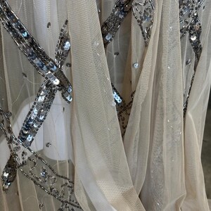 May include: Close-up of a sheer, beige fabric embellished with silver sequins. The sequins are arranged in a geometric pattern, creating a sparkling effect. The fabric is draped, showing folds and texture, suitable for evening wear.