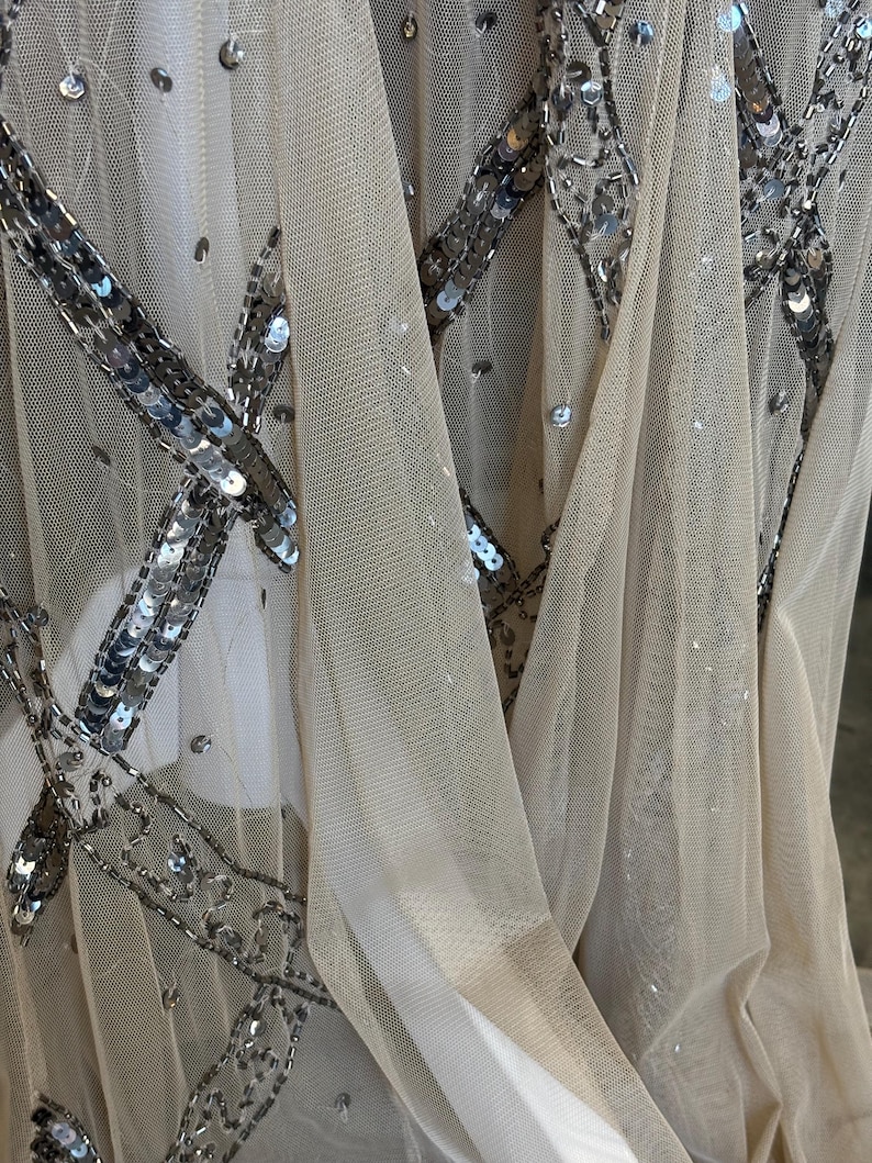 May include: Close-up of a sheer, beige fabric embellished with silver sequins. The sequins are arranged in a geometric pattern, creating a sparkling effect. The fabric is draped, showing folds and texture, suitable for evening wear.