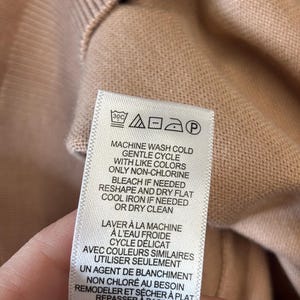 Puede incluir: Primer plano de una prenda de punto color melocot&oacute;n con una etiqueta de cuidado. La etiqueta proporciona instrucciones de lavado, incluyendo lavado a m&aacute;quina en fr&iacute;o, ciclo suave y planchado en fr&iacute;o si es necesario.