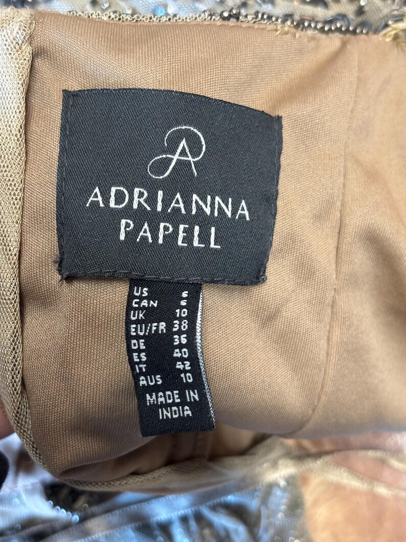 May include: A close-up of a black tag with the brand name "ADRIANNA PAPELL" in white lettering. The tag also includes size information for US, CAN, UK, EU/FR, DE, ES, IT, and AUS sizes. The tag reads "MADE IN INDIA".