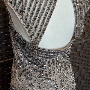 May include: A beige evening gown with intricate silver beadwork. The dress features a high neckline, cap sleeves, and an open back. The beading creates vertical and geometric patterns, adding texture and sparkle to the elegant design.