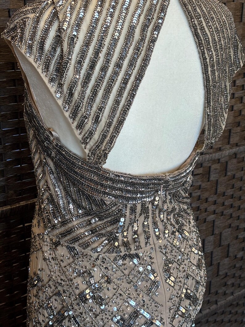 May include: A beige evening gown with intricate silver beadwork. The dress features a high neckline, cap sleeves, and an open back. The beading creates vertical and geometric patterns, adding texture and sparkle to the elegant design.