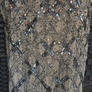 May include: Close-up of a beige dress with intricate silver sequin and bead embellishments. The design features a geometric pattern of intersecting lines and scattered sequins, creating a shimmering effect. The dress appears to be made of a sheer fabric.