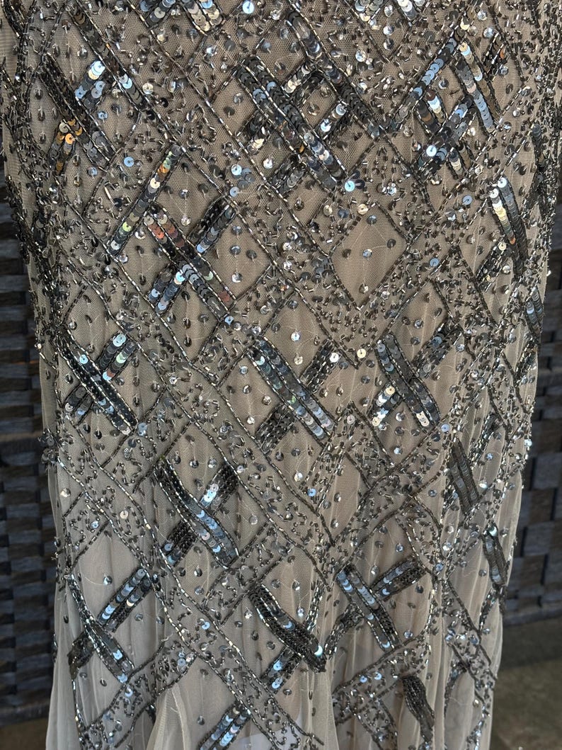 May include: Close-up of a beige dress with intricate silver sequin and bead embellishments. The design features a geometric pattern of intersecting lines and scattered sequins, creating a shimmering effect. The dress appears to be made of a sheer fabric.