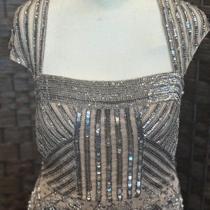 May include: A formal dress with a square neckline and cap sleeves. The dress is embellished with silver sequins and beads, creating a shimmering effect. The bodice features vertical and diagonal beaded patterns, adding texture and visual interest. The dress is displayed on a mannequin.