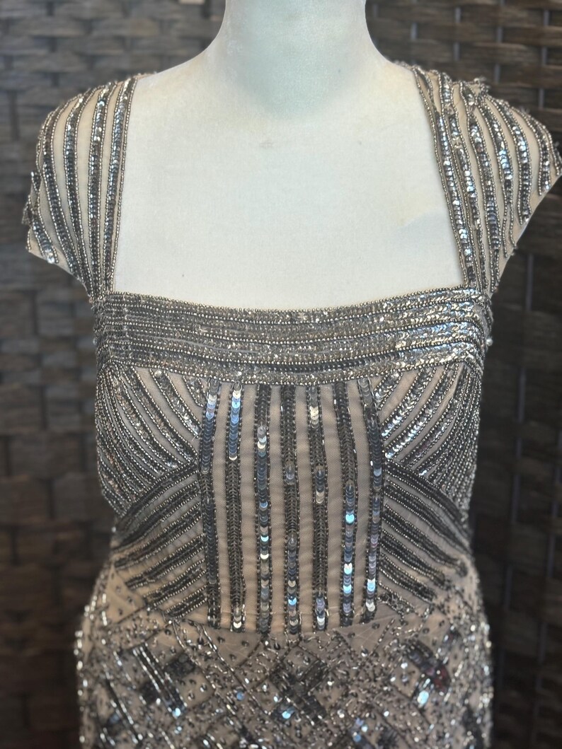 May include: A formal dress with a square neckline and cap sleeves. The dress is embellished with silver sequins and beads, creating a shimmering effect. The bodice features vertical and diagonal beaded patterns, adding texture and visual interest. The dress is displayed on a mannequin.