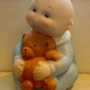 Op de afbeelding: Een keramieken beeldje van een baby die een teddybeer vasthoudt. De baby heeft een kale kop, blauwe ogen en draagt een lichtblauwe outfit. De teddybeer is bruin en wordt in de armen van de baby gehouden.