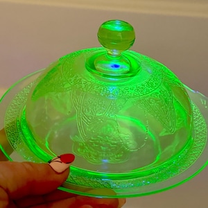 Könnte beinhalten: Eine leuchtend grüne Glasbutterdose mit gewölbtem Deckel und rundem Griff. Die Dose hat ein dekoratives Blumenmuster und einen gewellten Rand. Das Glas scheint Vintage zu sein und leuchtet hell.