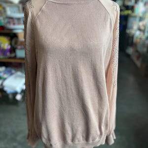 Puede incluir: Un su&eacute;ter rosa claro con cuello alto y mangas largas. Las mangas tienen paneles transparentes en los hombros, adornados con peque&ntilde;os adornos brillantes. El su&eacute;ter se exhibe en un maniqu&iacute;.