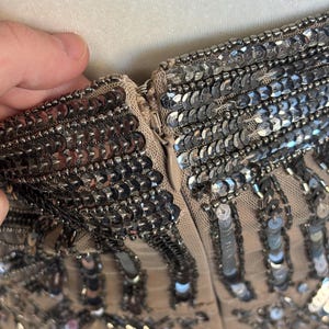 May include: Close-up of a beige garment with intricate silver sequin embellishments. The sequins are densely packed, creating a shimmering effect. A zipper is visible, and the fabric appears to be a delicate mesh.