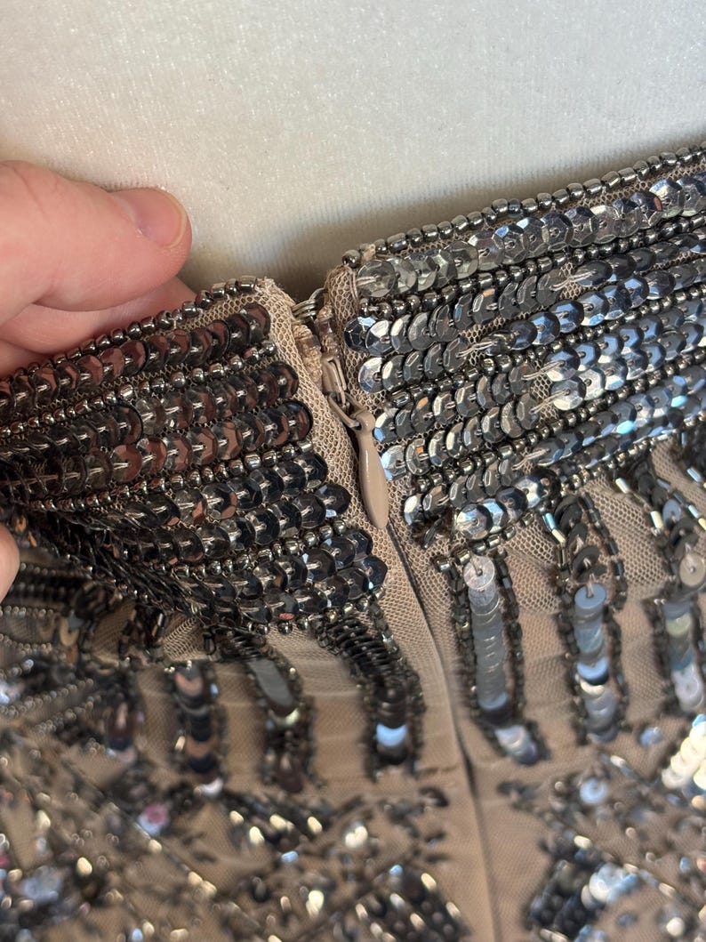 May include: Close-up of a beige garment with intricate silver sequin embellishments. The sequins are densely packed, creating a shimmering effect. A zipper is visible, and the fabric appears to be a delicate mesh.