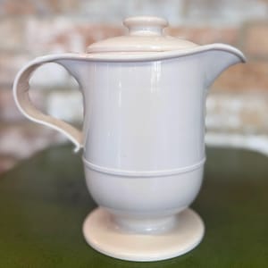 Peut inclure: Une cruche en céramique crème avec un couvercle, une poignée et un bec verseur. La cruche a un corps arrondi qui repose sur une base circulaire. La poignée s'incurve gracieusement du haut vers la base. L'arrière-plan est un mur de briques flou et une surface verte.