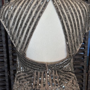 May include: A formal dress with a nude-colored base, adorned with intricate silver beading and sequins. The dress features a keyhole back and a detailed pattern, creating a glamorous and elegant look. The dress is displayed on a mannequin.