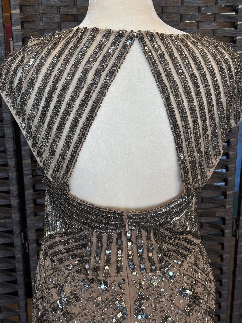 May include: A formal dress with a nude-colored base, adorned with intricate silver beading and sequins. The dress features a keyhole back and a detailed pattern, creating a glamorous and elegant look. The dress is displayed on a mannequin.