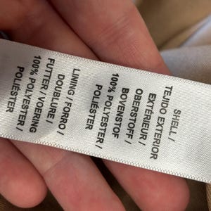May include: A close-up of a white clothing label with text in multiple languages, including English, Spanish, French, and German. The label lists the shell and lining materials as 100% polyester.