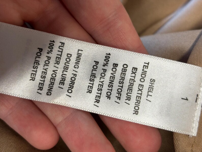 May include: A close-up of a white clothing label with text in multiple languages, including English, Spanish, French, and German. The label lists the shell and lining materials as 100% polyester.