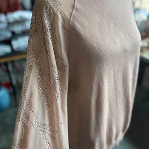 Puede incluir: Un su&eacute;ter marr&oacute;n claro con mangas transparentes. Las mangas est&aacute;n adornadas con brillantes brillantes. El su&eacute;ter tiene un cuello alto y una silueta ajustada. La prenda se exhibe en un maniqu&iacute;.