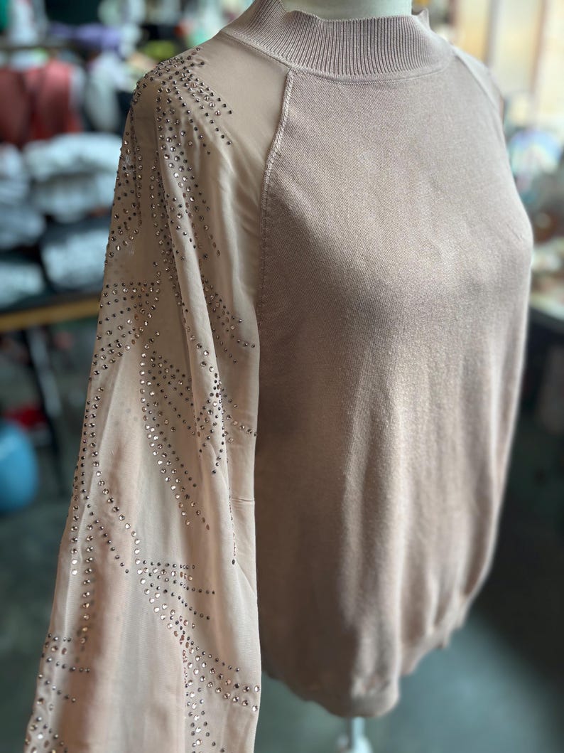 Puede incluir: Un su&eacute;ter marr&oacute;n claro con mangas transparentes. Las mangas est&aacute;n adornadas con brillantes brillantes. El su&eacute;ter tiene un cuello alto y una silueta ajustada. La prenda se exhibe en un maniqu&iacute;.
