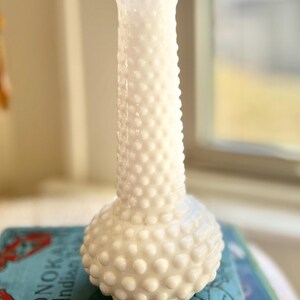 May include: A white hobnail vase with a bulbous base and a slender neck. The vase is made of opaque glass and has a textured surface with small, rounded bumps. The vase is sitting on top of a book.