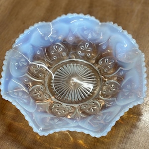 Puede incluir: Un pequeño plato de vidrio opalino con borde festoneado. El plato tiene un diseño de estrella en el centro y diseños florales alrededor del borde. El vidrio tiene un brillo suave e iridiscente, con toques de azul y melocotón.