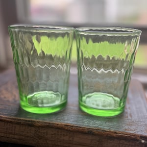 May include: Two vintage green drinking glasses with a textured, honeycomb-like pattern. The glasses are made of clear glass and have a vibrant, light green color. They are sitting on a dark, textured surface, likely a wooden table or shelf.