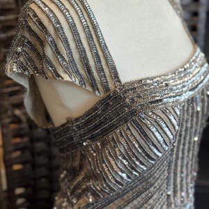 May include: Close-up of a beige evening gown with intricate silver sequin and bead embellishments. The dress features a square neckline and cap sleeves, with detailed patterns across the bodice and shoulders. The fabric appears to be a sheer mesh.