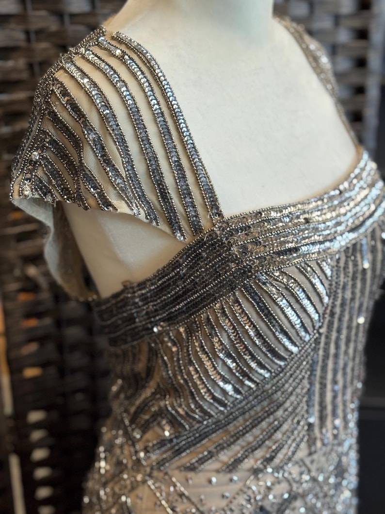 May include: Close-up of a beige evening gown with intricate silver sequin and bead embellishments. The dress features a square neckline and cap sleeves, with detailed patterns across the bodice and shoulders. The fabric appears to be a sheer mesh.