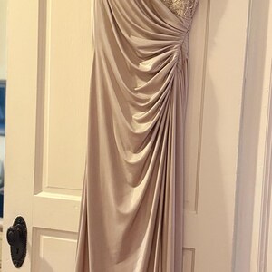 Puede incluir: Un vestido de noche largo, color champ&aacute;n, con un corpi&ntilde;o drapeado y una superposici&oacute;n de encaje con cuentas en la parte superior del pecho. El vestido cuelga de una percha de madera contra una puerta blanca.