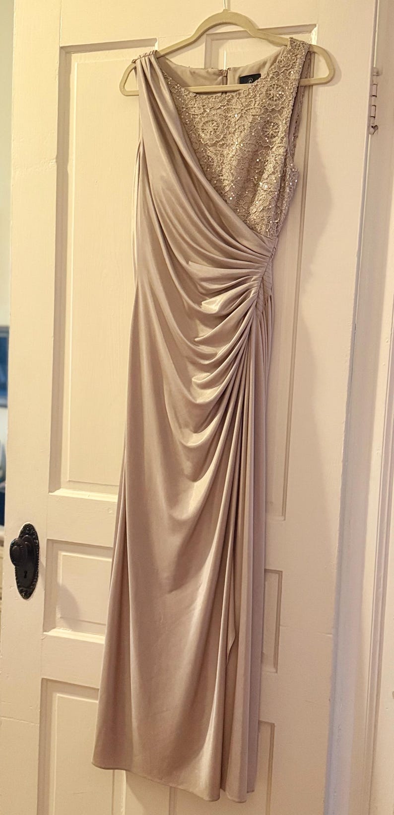 Puede incluir: Un vestido de noche largo, color champ&aacute;n, con un corpi&ntilde;o drapeado y una superposici&oacute;n de encaje con cuentas en la parte superior del pecho. El vestido cuelga de una percha de madera contra una puerta blanca.