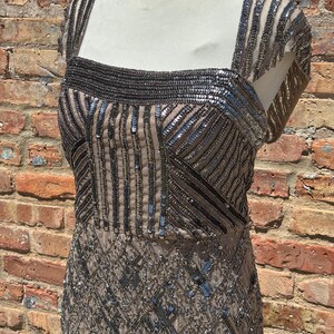 May include: A beige, sequined evening dress with a square neckline and cap sleeves. The bodice features vertical and horizontal sequin patterns, while the skirt has a diamond pattern. The dress is displayed on a mannequin against a brick wall.