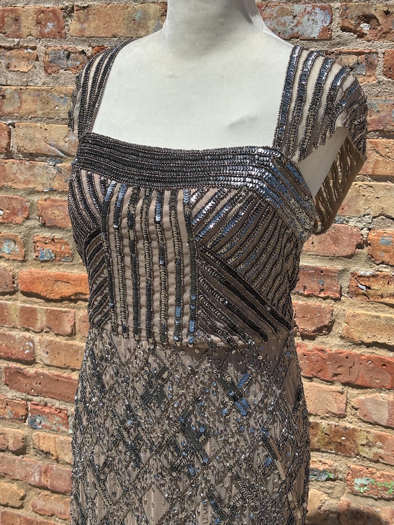 May include: A beige, sequined evening dress with a square neckline and cap sleeves. The bodice features vertical and horizontal sequin patterns, while the skirt has a diamond pattern. The dress is displayed on a mannequin against a brick wall.
